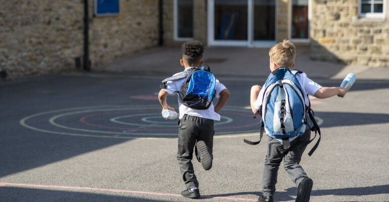 Rear view of two boys running in their school yard in the North East of England. They are running towards the door with their backpacks on.