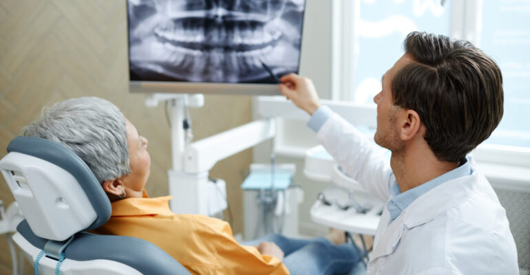 Back view of male dentist pointing at tooth X-ray image on screen during consultation in modern dental clinic
